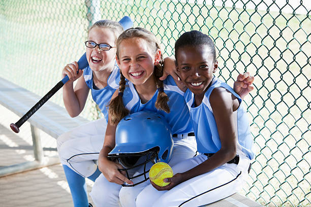An image of three girls with softball equipment sitting in a dugout.